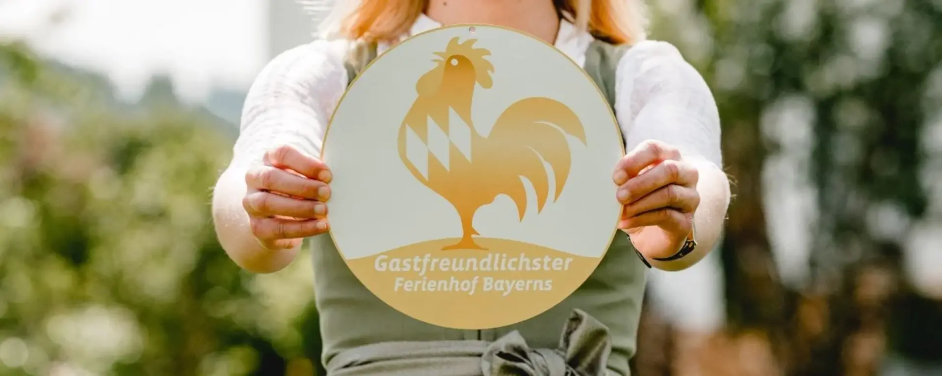 Gastgeberin mit Goldener Gockel Plakette - © Kimpelehof Eine Gastgeberin mit ihrer Plakette der Auszeichnung Goldener Gockel - Gastfreundlichster Ferienhof Bayerns im Dirndl im Garten ihres Urlaubsbauernhofes.