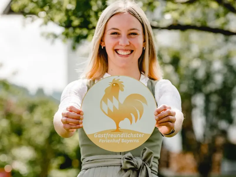 Gastgeberin mit Goldener Gockel Plakette - © Kimpelehof Eine Gastgeberin mit ihrer Plakette der Auszeichnung Goldener Gockel - Gastfreundlichster Ferienhof Bayerns im Dirndl im Garten ihres Urlaubsbauernhofes.